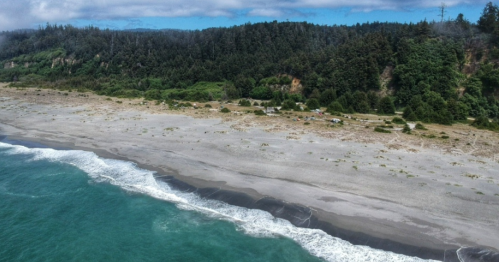 Aerial view of a sandy beach with gentle waves, surrounded by lush green forests and rocky cliffs in the background.