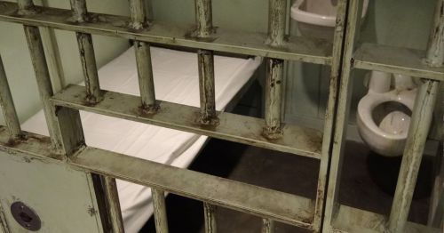 A view through prison bars showing a simple bed and a toilet in a small, dimly lit cell.