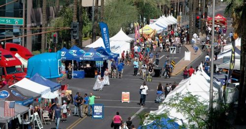 A bustling street fair with numerous tents, vendors, and crowds of people enjoying the event under sunny skies.