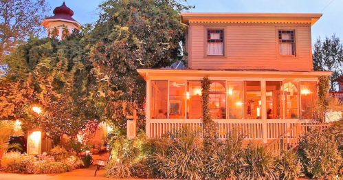 A charming two-story house with a porch, surrounded by lush greenery and warm evening lights.