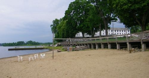 A sandy beach with white chairs, a wooden boardwalk, and trees along the shore under a cloudy sky.