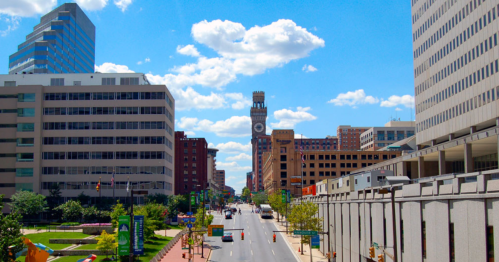 A city street lined with buildings under a bright blue sky with fluffy clouds, featuring a clock tower in the distance.
