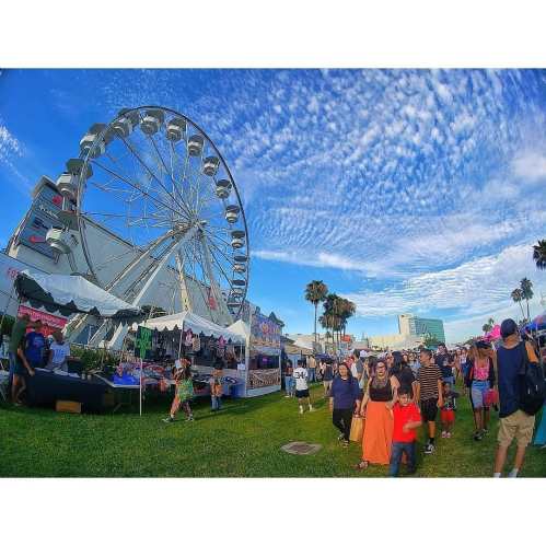 A lively fair scene with a large Ferris wheel, colorful tents, and families enjoying the festivities under a blue sky.