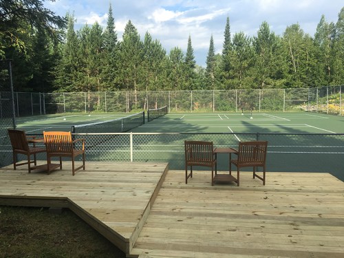A view of empty tennis courts surrounded by trees, with two wooden chairs on a deck in the foreground.