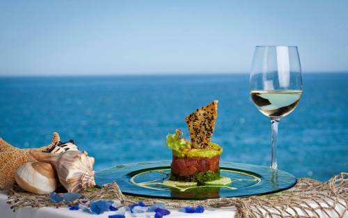 A gourmet dish with seafood and a decorative chip, accompanied by a glass of white wine, set against a seaside backdrop.