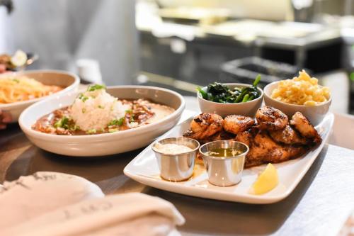 A plate of grilled chicken with sauces, sides of greens, macaroni, and a bowl of rice with beans in the background.