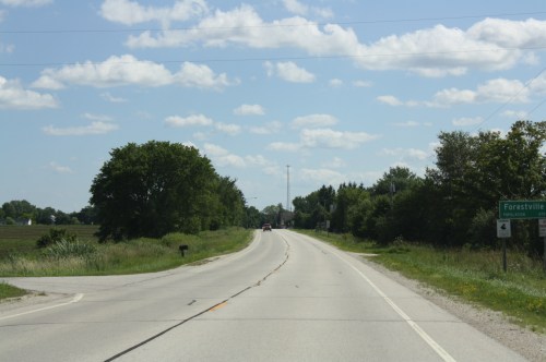 A rural road curves ahead, flanked by trees and fields, with a sign for Forestville visible on the right.