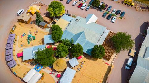 Aerial view of a recreational area with sandy play zones, trees, and parked cars. Various structures and amenities visible.
