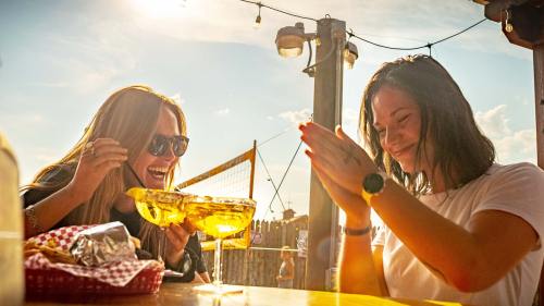 Two women enjoying a sunny outdoor meal, one laughing while holding a large drink and the other clapping joyfully.