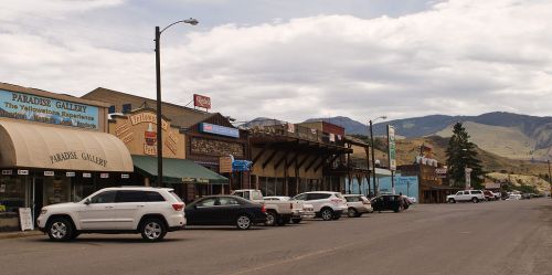 A small town street with shops, parked cars, and mountains in the background under a cloudy sky.
