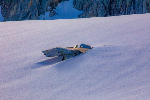A partially buried aircraft wreckage on a snowy glacier, surrounded by rugged mountain terrain.