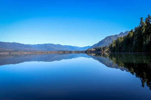 A serene lake reflecting mountains and a clear blue sky, surrounded by lush trees.