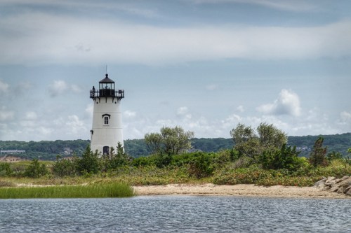 A white lighthouse stands near a calm water body, surrounded by greenery and under a cloudy sky.