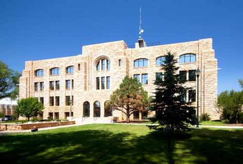 A large stone building with multiple windows, surrounded by green grass and trees under a clear blue sky.