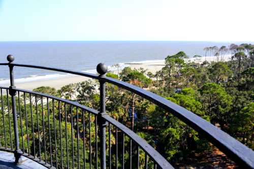 View from a balcony overlooking a sandy beach and ocean, framed by lush green trees under a clear blue sky.