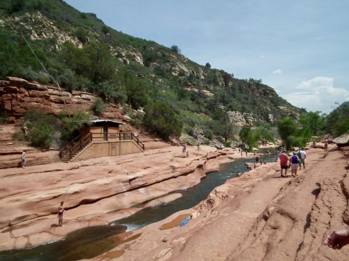 A scenic view of a rocky landscape with a stream, people walking, and lush greenery in the background.