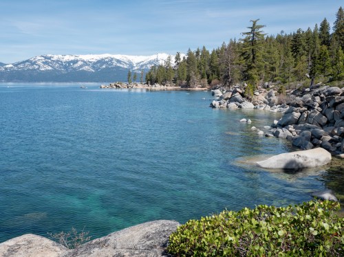 A serene lake scene with clear blue water, rocky shores, and snow-capped mountains in the background, surrounded by pine trees.