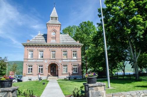 Historic brick building with a clock tower, surrounded by greenery and flowers under a clear blue sky.
