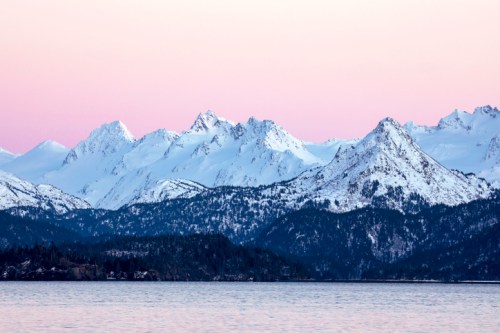 Snow-capped mountains rise above a calm lake, with a soft pastel sky at dawn.