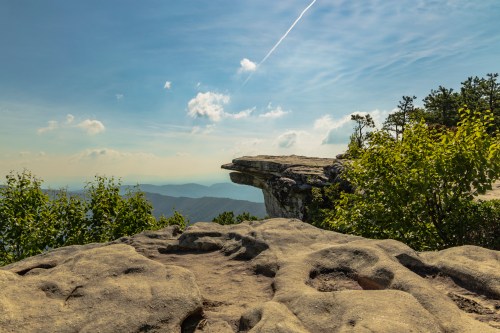 A rocky cliff edge overlooks a scenic landscape with distant mountains and a blue sky dotted with clouds.