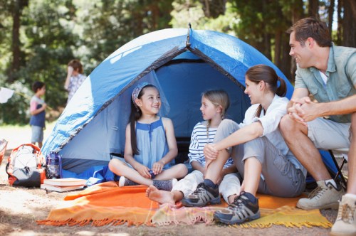 A family enjoys time together outside a blue tent, sitting on blankets and smiling in a forested campsite.