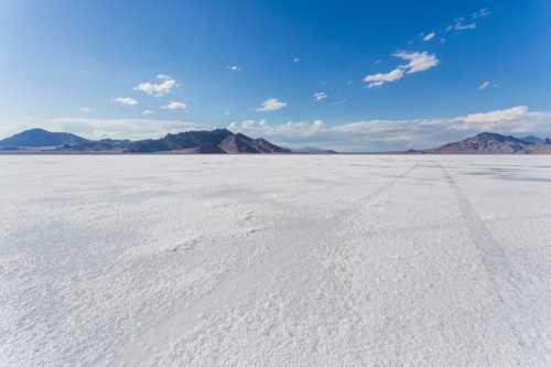 A vast, white salt flat stretches under a blue sky with distant mountains and scattered clouds.