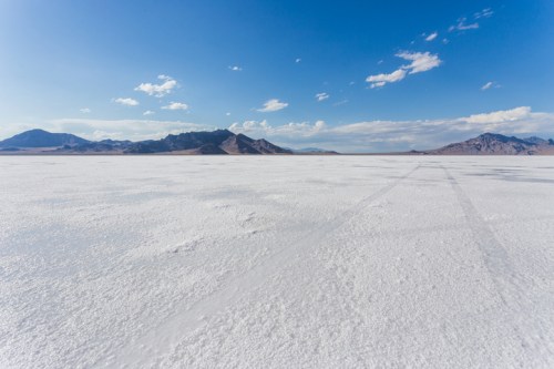 A vast, white salt flat stretches under a blue sky with distant mountains and scattered clouds.