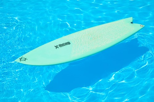 A light green surfboard floating in clear blue water, casting a shadow below.