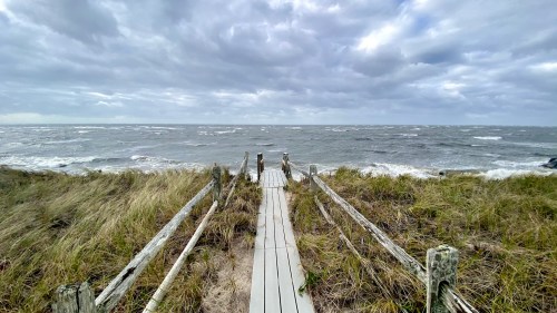 A wooden walkway leads to a stormy ocean, surrounded by tall grass under a cloudy sky.