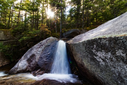 A serene waterfall cascades over rocks, surrounded by lush green trees and dappled sunlight filtering through the forest.