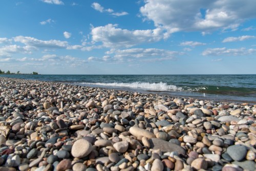 A rocky beach with smooth pebbles, gentle waves, and a blue sky dotted with fluffy clouds.