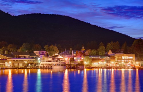 A serene lakeside view at dusk, with colorful lights reflecting on the water and a mountain backdrop.