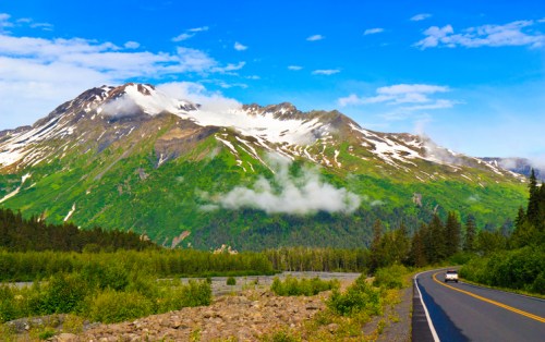 A scenic mountain landscape with snow-capped peaks, lush greenery, and a winding road under a blue sky with clouds.