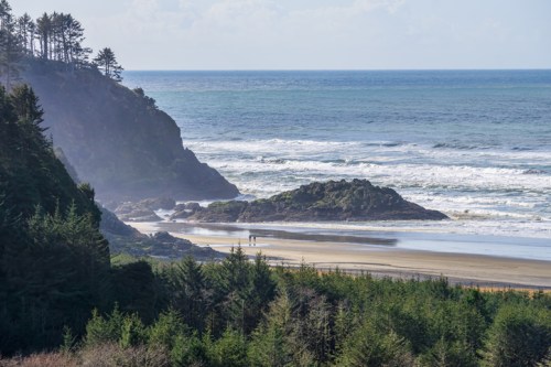 A scenic beach view with waves, rocky cliffs, and two people walking along the shore, surrounded by lush greenery.