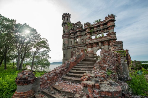 Ruins of a castle with crumbling walls and overgrown vegetation, set against a cloudy sky and a body of water.
