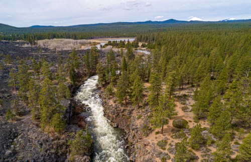 Aerial view of a winding river surrounded by lush green trees and rocky terrain, with distant mountains in the background.