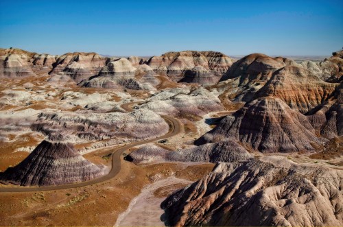 A winding road through colorful, layered rock formations in a vast desert landscape under a clear blue sky.