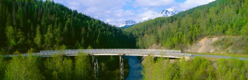 A scenic view of a bridge over a river, surrounded by lush green trees and snow-capped mountains in the background.