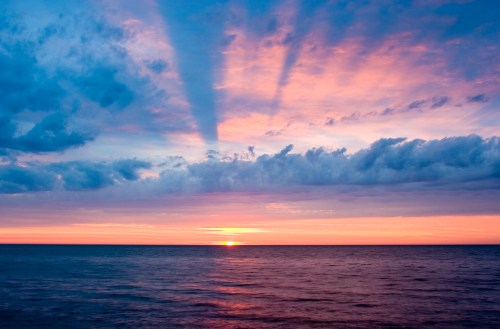 A serene sunset over the ocean, with vibrant colors and clouds casting long shadows across the water.