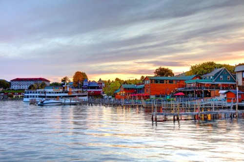 A scenic waterfront view featuring colorful buildings, boats, and a calm lake under a cloudy sunset sky.