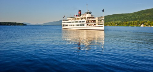 A large white steamboat sails on calm blue waters, surrounded by green mountains under a clear sky.