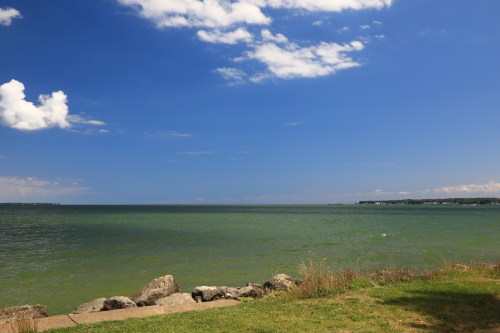 A serene view of a calm lake under a blue sky with scattered clouds, framed by grassy shore and rocky edge.