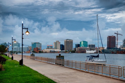 A waterfront view featuring a path, a sailboat, and a skyline with buildings under a cloudy sky.