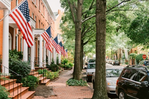 A tree-lined street with brick townhouses displaying American flags and parked cars along the curb.