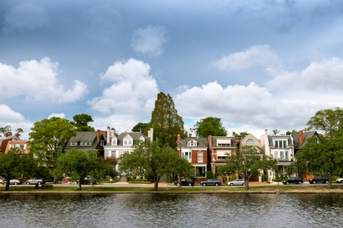 A scenic view of a row of houses along a river, with lush trees and a cloudy sky in the background.
