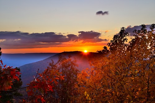 A vibrant sunset over a mountain, framed by colorful autumn leaves and a clear sky with scattered clouds.