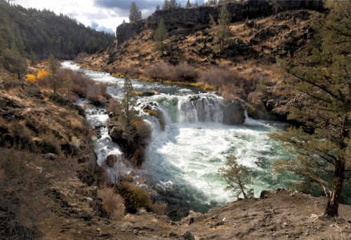 A scenic view of a waterfall cascading into a river, surrounded by rocky terrain and trees under a cloudy sky.