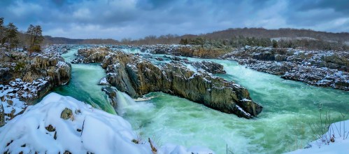 A snowy landscape featuring a river with rocky outcrops and flowing turquoise water under a cloudy sky.