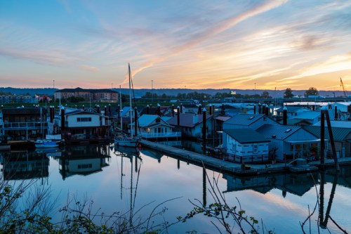 Sunset over a marina with houseboats and sailboats reflected in calm water, surrounded by hills and a colorful sky.