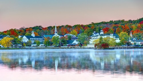 A serene lakeside view with colorful autumn trees and charming buildings reflected in the water at dawn.